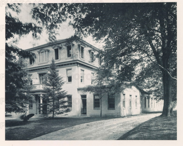 Department of steam engineering, photograph, page, photographer: Edward H. Hart, United States Naval Academy, cliché maker: The Photo-gravure Co., New York (city), c. 1885 - in or before 1890, North America, Maryland, Annapolis, paper, collotype, photoprint, height 183 mm x width 228 mm, machine driven by steam, facade (of house or building), military academy, A large multi-story brick building with a decorative upper facade and multiple windows is partially obscured by trees; a covered entrance with columns and a small balcony is visible on one side, and a dirt driveway runs along the front edge of the building; mature trees with dense foliage cast shadows across the scene and a metal object with curved blades sits on the lawn near the building.