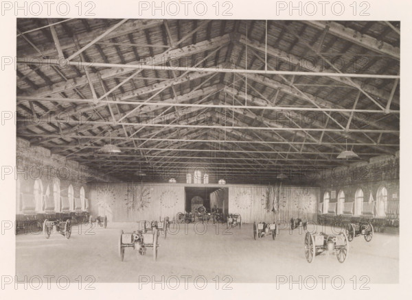 Armory, photograph, page, photographer: Edward H. Hart, United States Naval Academy, cliché maker: The Photo-gravure Co., New York (city), c. 1885 - in or before 1890, North America, Maryland, Annapolis, paper, collotype, photoprint, height 151 mm x width 219 mm, firearms: cannon, ammunition store, arsenal, A large interior space with a high wooden roof supported by exposed beams and trusses, hanging light fixtures suspended from the ceiling, brick side walls with a row of arched windows on each side, wooden flooring, a wide central doorway at the far wall flanked by decorative radial arrangements mounted on the wall, and several wheeled cannons or artillery pieces arranged across the floor.