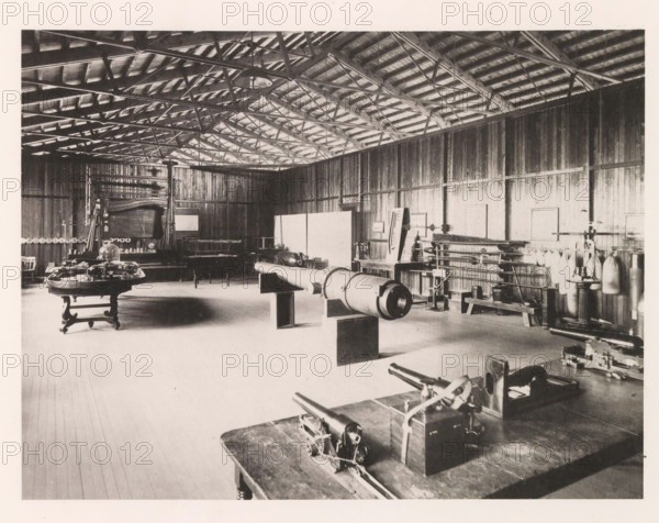 Torpedo model room, photograph, page, photographer: Edward H. Hart, United States Naval Academy, cliché maker: The Photo-gravure Co., New York (city), c. 1885 - in or before 1890, North America, Maryland, Annapolis, paper, collotype, photoprint, height 176 mm x width 234 mm, firearms: cannon, ammunition store, arsenal, A wide interior view of a large wooden room with exposed roof trusses and rafters, wooden plank walls and a wooden floor, containing various pieces of equipment and furniture arranged across the space, including a long cylindrical object mounted on supports near the center, several tables with mechanical devices on them in the foreground and left side, a tall framed apparatus against the back wall, shelving and racks holding cylindrical and bag-like items along the right wall, chairs and benches scattered around, and pendant lights hanging from the ceiling.