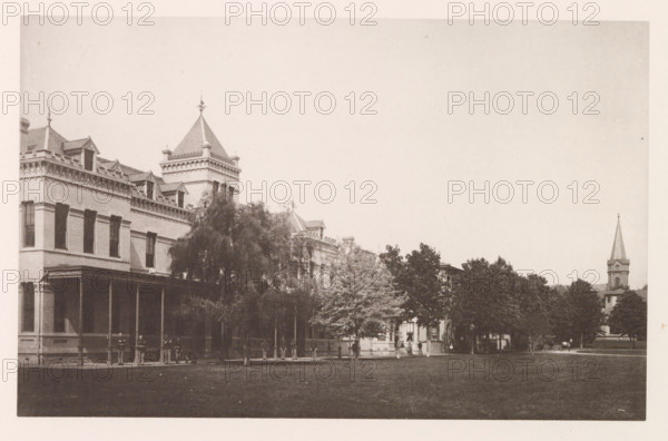 Porter Row, photograph, page, photographer: Edward H. Hart, United States Naval Academy, cliché maker: The Photo-gravure Co., New York (city), c. 1885 - in or before 1890, North America, Maryland, Annapolis, paper, collotype, photoprint, height 150 mm x width 237 mm, facade (of house or building), military academy, A wide sepia photograph showing the facade of a large brick building with decorative cornices, dormer windows, and a steep pyramidal tower topped with a finial, a covered porch with columns along its ground floor, and a row of trees partially obscuring the lower part of the building; several people stand or sit near the porch and along the sidewalk beneath the trees; a grassy lawn occupies the foreground, and to the right a church steeple rises above more trees with a small carriage visible near its base.