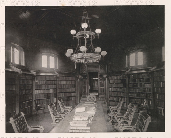 Chapel, photograph, page, photographer: Edward H. Hart, United States Naval Academy, cliché maker: The Photo-gravure Co., New York (city), c. 1885 - in or before 1890, North America, Maryland, Annapolis, paper, collotype, photoprint, height 177 mm x width 225 mm, public library, military academy, A high-ceilinged room with tall paneled bookshelves lining curved walls, several arched windows with light filtering through, a long central table covered with stacked books and papers, ornate upholstered chairs with carved wooden frames positioned along both sides of the table, a large multi-tiered chandelier hanging from the ceiling with spherical light fixtures and decorative chains, carved wooden columns and molding around the shelves, and a doorway visible at the far end leading to a darker interior space.