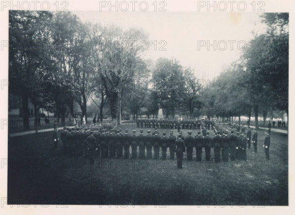 General muster, photograph, page, photographer: Edward H. Hart, United States Naval Academy, cliché maker: The Photo-gravure Co., New York (city), c. 1885 - in or before 1890, North America, Maryland, Annapolis, paper, collotype, photoprint, height 165 mm x width 240 mm, commander-in-chief, general, marshal, military academy, A wide outdoor scene showing a formal formation of uniformed individuals standing in concentric arcs on a grassy area surrounded by tall trees, with a central open space and an obelisk-like monument visible among the trees in the midground, pathways and scattered figures along the tree line, and a pale sky above.
