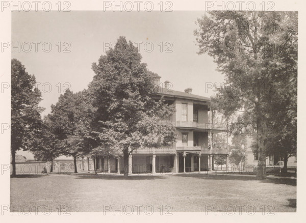 Sick-quarters, photograph, page, photographer: Edward H. Hart, United States Naval Academy, cliché maker: The Photo-gravure Co., New York (city), c. 1885 - in or before 1890, North America, Maryland, Annapolis, paper, collotype, photoprint, height 165 mm x width 236 mm, hospital, facade (of house or building), A large two-story building with a wraparound porch and ornate railing sits partially obscured by tall leafy trees in a grassy open area, with columns supporting the porch and a smaller covered entry at ground level; the building has tall windows and chimneys visible above the roof, and additional trees and parts of other brick structures are visible in the background.