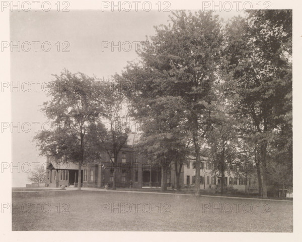 Superintendent's residence, photograph, page, photographer: Edward H. Hart, United States Naval Academy, cliché maker: The Photo-gravure Co., New York (city), c. 1885 - in or before 1890, Edward H. Hart, The Photo-gravure Co., photographer, cliché maker, North America, Maryland, Annapolis, paper, collotype, photoprint, height 185 mm x width 233 mm, housing, facade (of house or building), military academy, A wide lawn in the foreground leads to a row of large trees with dense canopies that dominate the middle of the image, partially obscuring a multi-story brick building with a porch, columns, and multiple windows; a smaller gabled entrance and a street lamp are visible near the building, and the scene is framed by a light border around the photograph.