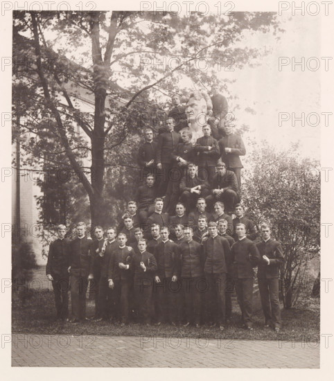 Class of '88, photograph, page, photographer: Edward H. Hart, United States Naval Academy, cliché maker: The Photo-gravure Co., New York (city), 1888, Edward H. Hart, The Photo-gravure Co., photographer, cliché maker, North America, Maryland, Annapolis, paper, collotype, photoprint, height 215 mm x width 185 mm, sculpture, officer, anonymous historical persons depicted in a group, in a group-portrait, A sepia-toned photograph shows a large group of young men arranged in rows outdoors, many wearing dark uniforms with high collars and holding hats or caps; some stand on the ground while others sit or perch higher, forming a pyramid shape around a large carved bust with a stern expression and stylized hair near the top center; behind them are tall trees with leafy branches and partial views of a building, and the foreground shows grass edging a paved surface.
