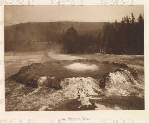 The Punch Bowl, page, photomechanical print, photographer: Frank Jay Haynes, Yellowstone National Park, cliché maker: The Photo-gravure Co., New York (city), c. 1882 - in or before 1887, North America, paper, photoaquatint, height 161 mm x width 213 mm, hot spring, geyser, A sepia-toned landscape showing a rocky, circular pool in the foreground with bubbling water at its center and mineral deposits around its rim, a textured ground with mineral streaks leading away from the pool, a middle ground of misty terrain with scattered trees and a fallen trunk, and a distant background of forested hills under a pale sky framed by a light border.