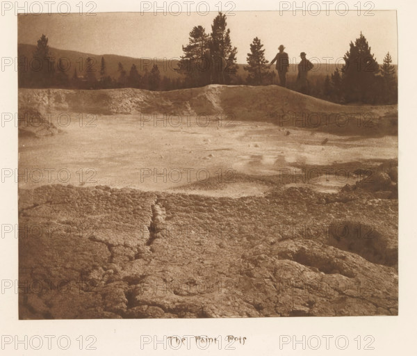 The paint pots, page, photomechanical print, photographer: Frank Jay Haynes, Yellowstone National Park, cliché maker: The Photo-gravure Co., New York (city), c. 1882 - in or before 1887, North America, paper, photoaquatint, height 170 mm x width 211 mm, pond, pool, hot spring, geyser, Fountain Paint Pot, A sepia-toned landscape photograph showing a rough, textured foreground of cracked and uneven ground leading to a flatter, lighter expanse and a low ridge; silhouettes of tall trees line the ridge against a pale sky, and three figures in dark clothing and hats stand near the ridge edge with two more distant tree silhouettes beyond.