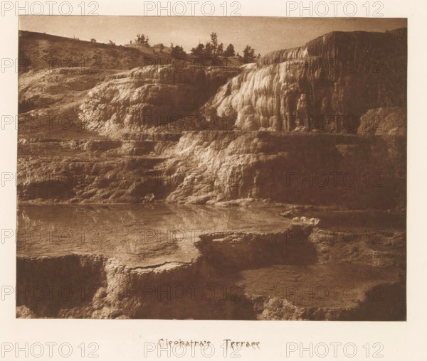 Cleopatra's terrace, page, photomechanical print, photographer: Frank Jay Haynes, Yellowstone National Park, cliché maker: The Photo-gravure Co., New York (city), c. 1882 - in or before 1887, Frank Jay Haynes, The Photo-gravure Co., photographer, cliché maker, North America, paper, photoaquatint, height 163 mm x width 209 mm, hot spring, geyser, A wide sepia-toned photograph showing a terraced travertine formation with layered, rounded ledges and cascading mineral textures rising from a flat pool at the bottom; the rock surfaces display striated, bulbous patterns and lighter highlights where the mineral deposits are thicker; a horizontal walkway or railing runs across one of the middle terraces; sparse trees and low vegetation line the ridge at the top against a smooth sky; the foreground includes water reflecting parts of the terraces and irregular mineral shelves projecting into the pool.