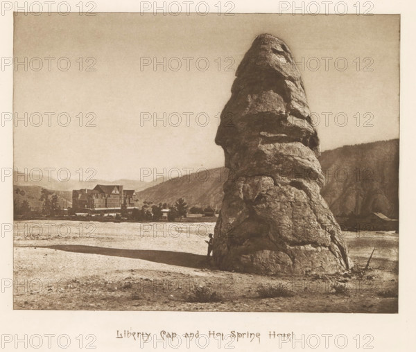Liberty Cap and hot spring hotel, page, photomechanical print, photographer: Frank Jay Haynes, Yellowstone National Park, cliché maker: The Photo-gravure Co., New York (city), c. 1882 - in or before 1887, North America, paper, photoaquatint, height 165 mm x width 215 mm, hotel, hostelry, inn, rock-formations, Liberty Cap (Yellowstone National Park), A large, tall, irregularly shaped rock formation dominates the right foreground, casting a long shadow across a flat, sandy plain; a small human figure stands at the rock's base, partially silhouetted against its textured surface while holding something; in the middle distance a multi-story building with steep gabled roofs and surrounding trees sits near the plain's edge with low hills rising behind it; distant ridges and a pale sky form the background.