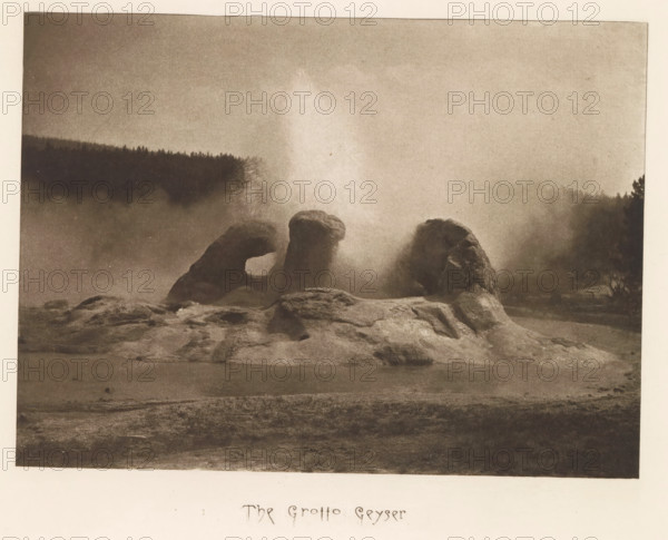The Grotto Geyser, page, photomechanical print, photographer: Frank Jay Haynes, Yellowstone National Park, cliché maker: The Photo-gravure Co., New York (city), c. 1882 - in or before 1887, North America, paper, photoaquatint, height 153 mm x width 210 mm, hot spring, geyser, Grotto Geyser, A sepia-toned landscape photograph showing a rocky formation rising from a flat foreground, with three rounded rock masses positioned close together and a tall plume of mist or steam erupting between them; a dark treeline stretches across the background beneath a hazy sky, and diffuse mist hangs over the midground creating soft, blurred edges around the rocks and distant trees.