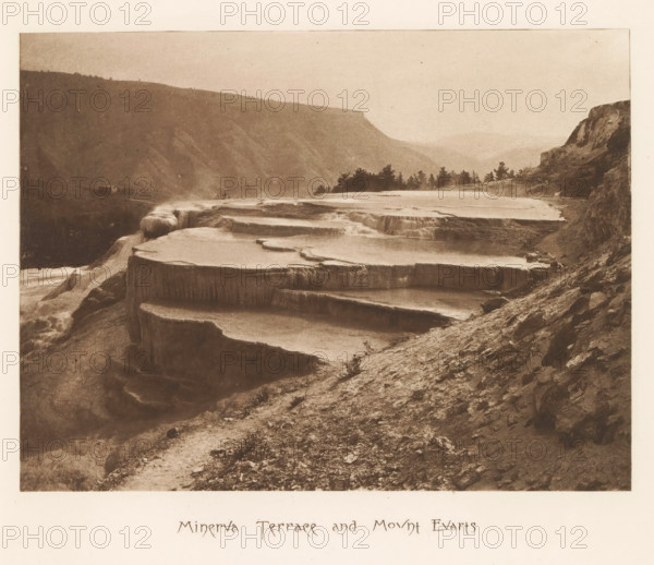 Minerva Terrace and Mount Evarts, page, photomechanical print, photographer: Frank Jay Haynes, Yellowstone National Park, cliché maker: The Photo-gravure Co., New York (city), c. 1882 - in or before 1887, North America, paper, photoaquatint, height 162 mm x width 216 mm, mountains, hot spring, geyser, Mount Everts, A sepia-toned landscape showing a series of broad terraced rock formations filled with shallow water cascading between levels, a steep rocky slope in the foreground with scattered small stones and sparse low vegetation, a distant ridge of hills fading into the background, a cluster of trees along the far terrace edge, and rocky cliffs rising on the right side of the image.