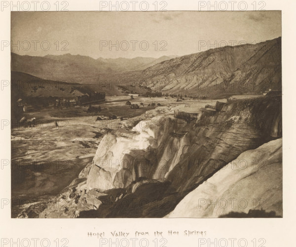 Hotel valley from the hot springs, page, photomechanical print, photographer: Frank Jay Haynes, Yellowstone National Park, cliché maker: The Photo-gravure Co., New York (city), c. 1882 - in or before 1887, North America, paper, photoaquatint, height 162 mm x width 216 mm, dale, valley, A wide sepia-toned landscape photograph showing a rocky foreground with terraces and smooth rock formations leading down toward a flat valley where small buildings and clustered trees are visible, beyond which low rolling hills and a range of rugged mountains extend across the background under a pale sky.