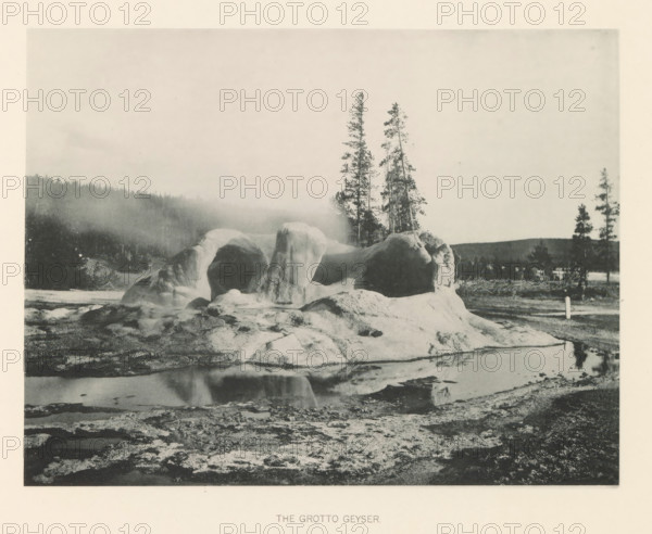 The Grotto Geyser, page, photomechanical print, photographer: Frank Jay Haynes, Yellowstone National Park, cliché maker: The Photo-gravure Co., New York (city), c. 1886 - in or before 1891, North America, paper, collotype, photoprint, height 165 mm x width 215 mm, hot spring, geyser, Grotto Geyser, A grayscale landscape shows a rounded rocky formation with smooth, bulbous surfaces and arch-like openings sitting beside a reflective pool of water, with the rock reflecting in the pool; behind the formation are tall, slender evergreen trees and a distant tree-covered hillside partially obscured by mist, while the foreground consists of rough, textured ground and patches of shallow water.
