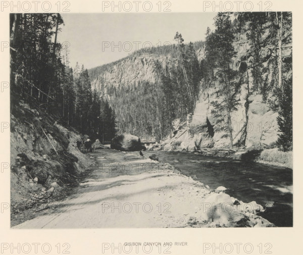 Gibbon Canyon and river, page, photomechanical print, photographer: Frank Jay Haynes, Yellowstone National Park, cliché maker: The Photo-gravure Co., New York (city), c. 1886 - in or before 1891, North America, paper, collotype, photoprint, height 163 mm x width 216 mm, river, Gibbon River, A black and white landscape showing a dirt road running alongside a flowing river with rocks and boulders at the water's edge, steep rocky cliffs and tall trees rising on the right bank, a densely forested slope and rugged cliff face in the background, a wooded hillside on the left with trees extending toward the sky, scattered rocks and tree roots along the roadside, and a small wooden bridge crossing the river near a large boulder.