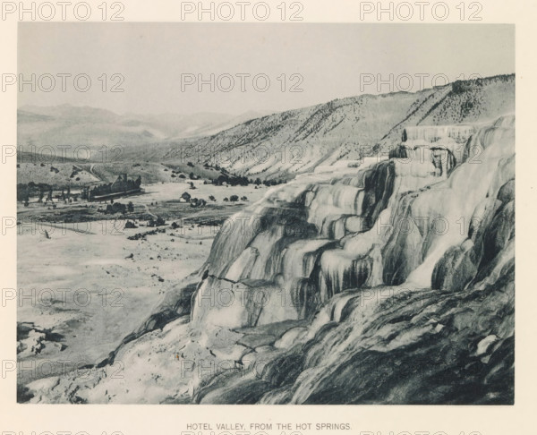 Hotel valley, from the hot springs, page, photomechanical print, photographer: Frank Jay Haynes, Yellowstone National Park, cliché maker: The Photo-gravure Co., New York (city), c. 1886 - in or before 1891, North America, paper, collotype, photoprint, height 165 mm x width 215 mm, dale, valley, hot spring, geyser, A wide landscape shows tiered mineral terraces with smooth rounded ledges and cascading surfaces in the foreground, their textures ranging from light to dark tones; beyond the terraces a flat valley contains scattered small buildings and a larger complex with peaked roofs, surrounded by open land and sparse trees; rolling hills and ridges rise in the background with scattered vegetation on their slopes, and a pale sky fills the upper portion of the scene.