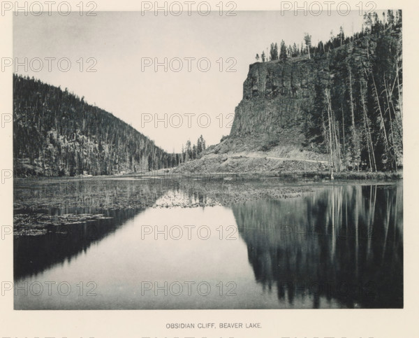 Obsidian cliff, Beaver Lake, page, photomechanical print, photographer: Frank Jay Haynes, Yellowstone National Park, cliché maker: The Photo-gravure Co., New York (city), c. 1886 - in or before 1891, North America, paper, collotype, photoprint, height 165 mm x width 215 mm, ravine, chasm, abyss, canyon, A wide black-and-white photograph shows a calm lake in the foreground reflecting the cliff and trees along its shore, with the water surface dotted with patches of vegetation and subtle ripples; to the right a tall rocky cliff rises from a sloping shoreline, topped by a line of trees and bordered by a stand of tall, thin trunks along the water's edge, while to the left a densely forested hillside slopes down to meet the shore, and a pale sky fills the upper area above the landscape.