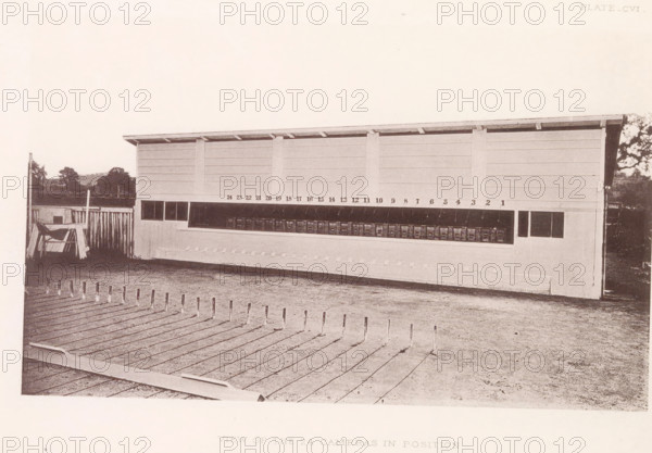 View of the 24 cameras in position, Setup with photo cameras to capture a horse in motion, page, photomechanical print, number: 'Plate_CVI.', photographer: Eadweard Muybridge, Stanford, cliché maker: anonymous, c. 1878 - in or before 1882, clichémaker, paper, photoaquatint, height 133 mm x width 217 mm, taking photographs - CC - out of doors, photographic apparatus, A long rectangular wooden structure with a slanted roof stands horizontally across the scene; its front face has a series of dark rectangular openings along a midline and a row of small square compartments beneath a sheltered overhang, with vertical supports dividing the panels. In front of the structure is an open yard with a boarded surface in the foreground featuring evenly spaced slender upright posts, and a patch of bare ground between the boards and the building. To the left of the structure there is a fenced area with a simple wooden frame and leaning planks, and low distant trees and shrubbery are visible along the ...