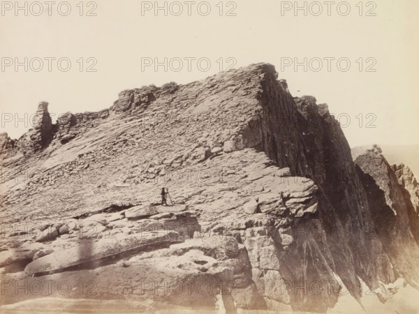 View of summit of Mount Hoffmann in the Yosemite Valley, Summit of Mt. Hoffmann, photograph, page, number: 'XXV.', photographer: W. Harris, Yosemite Valley, c. 1860 - in or before 1868, North America, California, paper, albumen print, height 157 mm x width 209 mm, mountain-top, peak, Mount Hoffmann, A sepia-toned photograph of a rocky mountain ridge with layered, jagged rock formations extending toward a steep cliff face, scattered boulders and slabs on the sloping foreground, a lone figure standing near a tripod on the rocky surface, and a tall, narrow rock outcrop visible farther along the ridge against a pale sky.