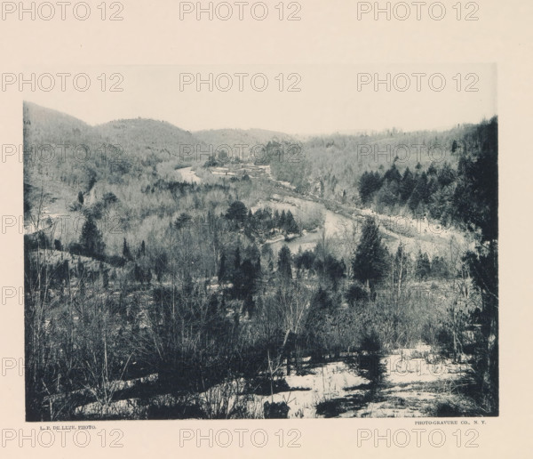 Site of proposed Quaker Bridge Dam and reservoir, page, photomechanical print, photographer: P. de Luze, New York, cliché maker: The Photo-gravure Co., c. 1883 - in or before 1887, North America, paper, collotype, photoprint, height 153 mm x width 202 mm, river, Croton, A wide, desaturated landscape photograph shows a river winding through a valley surrounded by forested hills, with bare trees and evergreens visible on the slopes and along the riverbanks, patches of snow on the ground in the foreground, denser dark trees on one side of the valley, and distant rolling hills fading toward the horizon.