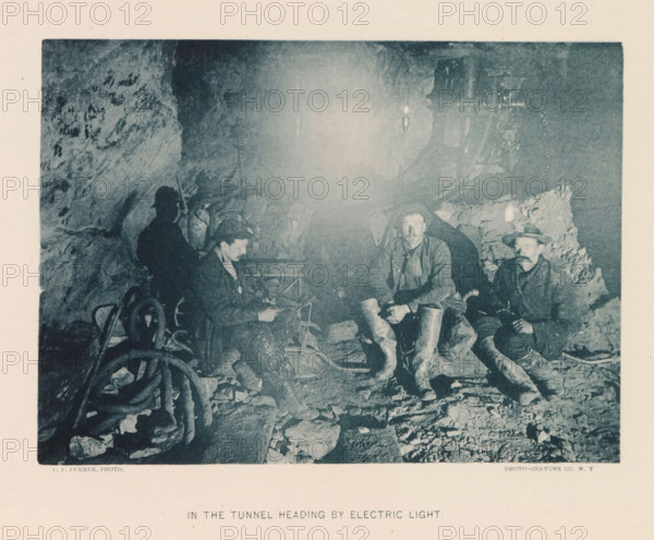 In the tunnel heading by electric light, page, photomechanical print, photographer: CF Ferrer, New York, cliché maker: The Photo-gravure Co., c. 1883 - in or before 1887, North America, paper, collotype, photoprint, height 142 mm x width 194 mm, underground road, tunnel, working class, laborers, A grainy monochrome photograph showing the interior of a rough rock tunnel where several men sit and crouch among stones and equipment; three men are visible in the foreground wearing work clothes and boots, one holding an object and another wearing a hat and looking forward, while a shadowed figure sits against the tunnel wall to the left; coiled hoses or cables and scattered rocks lie on the tunnel floor, and a bright light source creates a central glow that illuminates the rocky surfaces and casts shadows on the men and equipment.