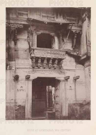 Façade of a house in Ahmedabad, Gujarat, India, page, photomechanical print, number: 'I.', title: 'HOUSE AT AHMEDABAD, 16TH CENTURY.', Ahmedabad (city), maker: Heliotype Printing Company, Boston (Massachusetts), c. 1875 - in or before 1885, photographer, North America, Massachusetts, paper, photoaquatint, height 230 mm x width 173 mm, urban housing, facade (of house or building), A sepia-toned photograph of a building facade showing an ornate balcony with carved pillars and decorative brackets above a large arched doorway. The balcony features intricate latticework panels and a row of sculpted corbels beneath its ledge. Flanking the doorway are weathered stone columns and remnants of carved details, with visible cracks and surface wear on the walls. Above the balcony is a wooden overhang with exposed beams and small balustrades along the upper level. The lower portion of the image shows the dark interior beyond the doorway and rough masonry at the base of the wall.