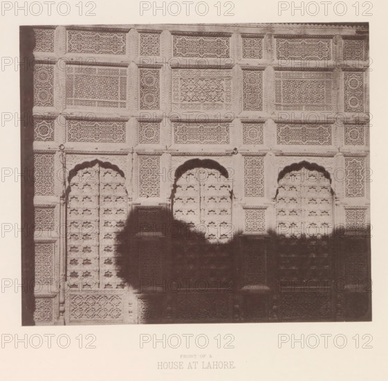Façade of a house in Lahore, Punjab, Pakistan, page, photomechanical print, number: 'XI.', title: 'FRONT OF A, HOUSE AT LAHORE.', Lahore, maker: Heliotype Printing Company, Boston (Massachusetts), c. 1875 - in or before 1885, photographer, North America, Massachusetts, paper, photoaquatint, height 169 mm x width 198 mm, urban housing, facade (of house or building), A sepia photograph shows a large carved wooden or stone façade with multiple rectangular panels filled with intricate latticework and geometric patterns, framed by raised borders and carved vertical and horizontal beams; three arched recessed doorways with decorative molding are visible in the middle portion, each door set with repeating ornamental motifs, and shadowed areas obscure parts of the lower central section; the surface displays varied textures from finely pierced screens to carved relief panels, and the image is bordered by a plain margin.