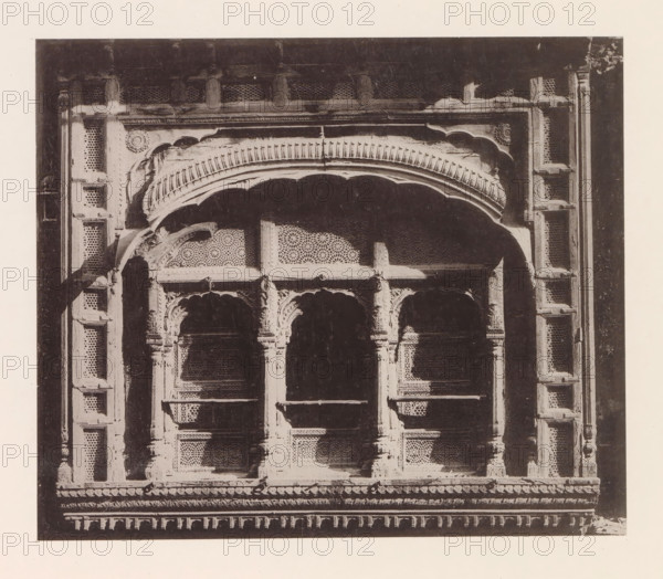 Façade of a house in Lahore, Punjab, Pakistan, page, photomechanical print, title: 'FRONT OF A, HOUSE AT LAHORE.', number: 'XII.', Lahore, maker: Heliotype Printing Company, Boston (Massachusetts), c. 1875 - in or before 1885, photographer, North America, Massachusetts, paper, photoaquatint, height 161 mm x width 190 mm, urban housing, façade (of house or building), A close-up view of an ornate stone or wood window facade with carved arches and decorative latticework panels, showing scalloped arch moldings and a prominent curved cornice above the openings, fluted pilasters and column-like supports with detailed capitals, repeated geometric perforated screens behind the arches, and a horizontal decorative ledge and molding at the base, all rendered in a high-contrast monochrome tone against a plain border.