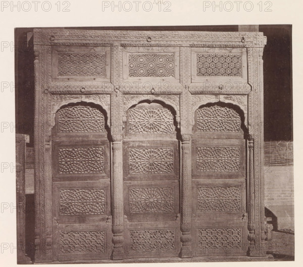 Window with geometrical ornaments in Lahore, Punjab, Pakistan, page, photomechanical print, title: 'WINDOW OF A, HOUSE AT LAHORE.', number: 'XIII.', Lahore, maker: Heliotype Printing Company, Boston (Massachusetts), c. 1875 - in or before 1885, photographer, North America, Massachusetts, paper, photoaquatint, height 177 mm x width 207 mm, ornament, geometric motifs, window, A carved wooden panel or door divided into vertical sections with repeating geometric lattice panels and ornate floral and rope-like border carvings, featuring arched recessed tops above central panels and fluted columns between sections, all shown in a monochrome photograph with a dark background and a light margin around the image.