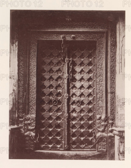 Door of a house in Multan, Punjab, Pakistan, page, photomechanical print, number: 'XVI.', title: 'DOOR OF HOUSE, MULTAN.', Multan, maker: Heliotype Printing Company, Boston (Massachusetts), c. 1875 - in or before 1885, photographer, North America, Massachusetts, paper, photoaquatint, height 221 mm x width 170 mm, closed door, A close view of an ornate wooden double door with a patterned grid of raised square studs and round metal ring handles, each door edged with carved vertical panels and a decorative central vertical strip; heavy metal chains hang across the doors and loop through anchors, and the surrounding frame is richly carved with floral and foliate motifs extending into the adjacent wall surfaces.