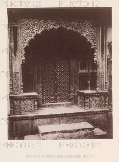 View of a doorway of a house in Multan, Punjab, Pakistan, page, photomechanical print, title: 'DOORWAY OF HOUSE, RED SANDSTONE, MULTAN.', number: 'XVIII.', Multan, maker: Heliotype Printing Company, Boston (Massachusetts), c. 1875 - in or before 1885, photographer, North America, Massachusetts, paper, photoaquatint, height 214 mm x width 170 mm, gate, entrance, closed door, A sepia-toned photograph of an ornate stone doorway featuring a scalloped arch with dense floral and vine carvings along its border, flanked by carved pilasters and panels with repeating floral motifs, a heavily decorated pair of wooden doors with geometric and floral relief patterns, raised stone platforms or benches on either side of the entrance with carved front panels, a low stone step leading up to the doorway, and weathered stone surfaces throughout.