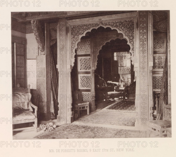 Interior of the house of Lockwood de Forest in New York, page, photomechanical print, number: 'XIX.', title: 'MR. THE FOREST'S ROOMS, 9 EAST 17TH ST., NEW YORK.', New York (city), maker: Heliotype Printing Company, Boston (Massachusetts), c. 1875 - in or before 1885, Heliotype Printing Company, photographer, North America, Massachusetts, paper, photoaquatint, height 185 mm x width 231 mm, interior of the house, decorations (interior of the house), A sepia photograph of an interior showing an ornately carved archway with scalloped edges and intricate floral patterns, flanked by carved pillars and panels with latticework screens visible behind the arch; patterned draperies hang at the sides and carved wooden furniture including upholstered chairs and small stools occupy the foreground and interior space; multiple patterned rugs cover the floor and a small decorative object lies near the front chair; light and shadow reveal textured surfaces and detailed woodwork throughout the room.
