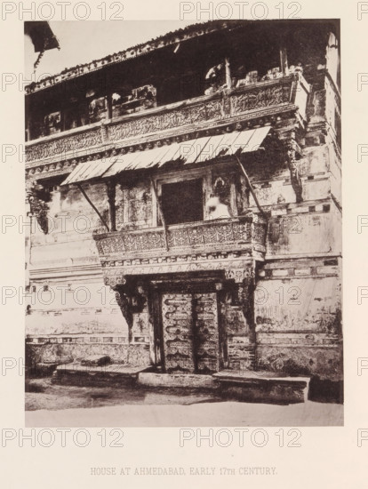 Façade of a house with a balcony in Ahmedabad, Gujarat, India, page, photomechanical print, title: 'HOUSE AT AHMEDABAD, EARLY 17TH CENTURY.', number: 'XXII.', Ahmedabad (city), maker: Heliotype Printing Company, Boston (Massachusetts), c. 1875 - in or before 1885, photographer, North America, Massachusetts, paper, photoaquatint, height 233 mm x width 182 mm, urban housing, balcony, façade (of house or building), A monochrome photograph of an ornate building façade showing a carved wooden balcony with decorative panels and a slanted awning supported by angled beams, an open window or doorway behind the balcony with a blurred light-toned figure or object visible inside, elaborately carved pillars and brackets beneath the balcony, a pair of intricately carved double doors at street level framed by decorative lintels, worn stone steps and a narrow pavement in front, weathered plastered walls with horizontal decorative bands, and an upper storey with a long carved frieze and a projecting ro...