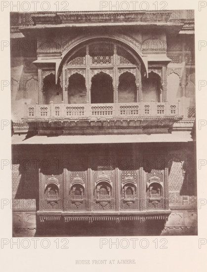 Façade of a house with a balcony in Ajmer, Rajasthan, India, page, photomechanical print, title: 'HOUSE FRONT AT AJMERE.', number: 'VI.', Ajmer, maker: Heliotype Printing Company, Boston (Massachusetts), c. 1875 - in or before 1885, photographer, North America, Massachusetts, paper, photoaquatint, height 231 mm x width 189 mm, urban housing, balcony, façade (of house or building), A sepia-toned photograph of a building facade showing ornate carved stonework and a projecting balcony with patterned balustrades and arched openings. The upper section features a large central arch flanked by smaller arches, each with decorative latticework and scalloped arch details, supported by slender columns with intricate carvings. Below the balcony is a row of elaborately carved panels with recessed arched niches and geometric and floral motifs. The stone surfaces display textured patterns and ornamental friezes, with shadows creating contrast across the architectural details.