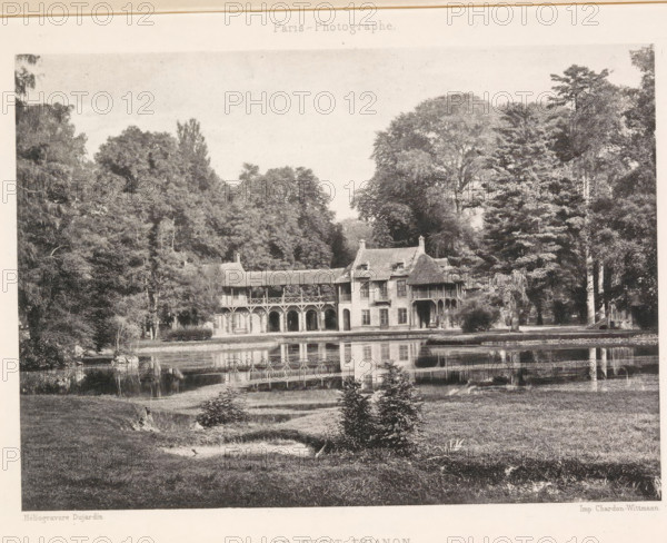 View of the Hameau de la Reine, Le Petit Trianon, page, photomechanical print, photographer: Dujardin, Versailles, maker: Chardon-Wittmann, c. 1881 - in or before 1891, paper, photoaquatint, height 122 mm x width 172 mm, palace, Le Hameau de la Reine, A monochrome image showing a large two-story building with arched openings on the ground level, a covered balcony supported by wooden posts on the upper level, and a steeply pitched roof with chimneys, set at the edge of a reflective body of water; dense trees of varying heights and shapes surround the building on both sides, and the water in front of the building mirrors its facade and the trees, with a grassy foreground containing small shrubs and a gently sloping bank.