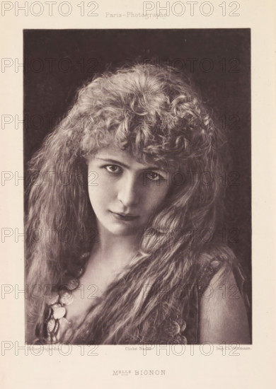 Melle Bignon, Portrait of Louise Bignon, page, photomechanical print, photographer: Félix Nadar, Paris, cliché maker: Dujardin, France, printer (person): Charles Wittmann, c. 1884 - in or before 1894, printer (person), paper, photoaquatint, height 155 mm x width 111 mm, Person, Louise Bignon, portrait of actor, actress, A sepia-toned photographic portrait of a young woman framed by a dark rectangular background, showing her head and upper torso; she has long, wavy hair with a voluminous, curly fringe and soft waves cascading over her shoulders, visible skin tones of a pale complexion, large eyes looking slightly upward, a straight nose, and closed lips with a faint neutral expression; she wears a garment with textured fabric and a necklace made of round, reflective discs that rest on her chest, and the image is bordered by a lighter margin.