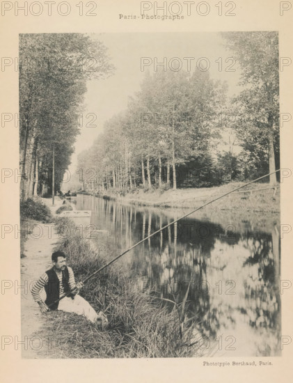 Aulnay-Les-Bois, Fisherman in Aulnay-sous-Bois, page, photomechanical print, photographer: anonymous, Aulnay-sous-Bois, cliché maker: Michel Berthaud, France, c. 1884 - in or before 1894, paper, collotype, photoprint, height 150 mm x width 120 mm, fisherman, A sepia-toned photograph showing a tree-lined canal with tall trees along both banks and their reflections in the water, a narrow dirt path beside the canal, a man sitting on the grass at the water’s edge holding a long fishing rod, grassy vegetation along the bank, and a small boat moored further along the canal.