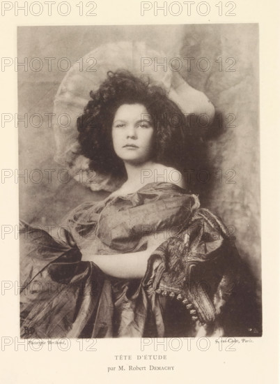 Tête d'étude, Portrait of an unknown woman with a hat, page, photomechanical print, photographer: Robert Demachy, France, cliché maker: Michel Berthaud, Paris, c. 1884 - in or before 1894, Robert Demachy, Michel Berthaud, photographer, cliché maker, paper, collotype, photoprint, height 153 mm x width 118 mm, adult woman, A sepia-toned portrait of a woman seated and facing forward with a neutral expression, her dark curly hair forming a voluminous halo around her head and a light, rounded fabric piece behind it; she wears an off-the-shoulder dress with gathered, slightly wrinkled fabric and a richly patterned draped textile over one arm, the visible arm resting across her lap, while the background shows a soft, mottled texture.