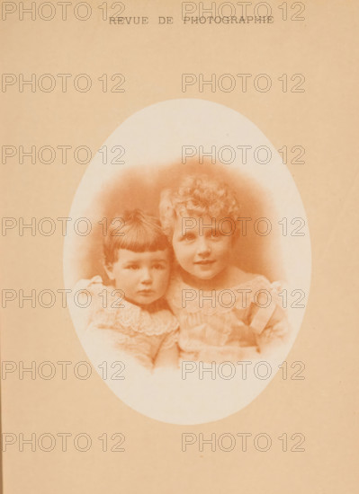 Portrait of two young unknown children, page, photomechanical print, inscription: 'Revue de Photographie', maker: F. Thévoz & Cie., c. 1887 - in or before 1892, paper, collotype, photoprint, height 120 mm x width 92 mm, historical persons, toddler, A faded oval sepia photograph of two young children framed by a blank textured background, showing a child with short straight hair and a lace-collared dress leaning against a child with curly hair wearing a frilled dress, both seated close together with neutral expressions and soft lighting that mutes facial details.