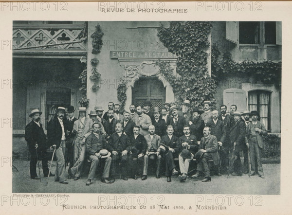 Group portrait of unknown men, Réunion photographique de 29 Mai 1892 a Monnetier, page, photomechanical print, inscription: 'Revue de Photographie', maker: Antoine Elie Chevalley, 1892-05-29, paper, collotype, photoprint, height 115 mm x width 179 mm, anonymous historical persons depicted in a group, in a group portrait, A large group of men posed in front of a building façade covered in climbing ivy and decorative stonework, many wearing suits and hats and some holding walking sticks, with a carved doorway centered behind them and shuttered windows visible to the sides, a patterned balcony railing at the upper left, and a bench along which several seated men are arranged while others stand around them.