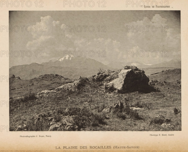 View of a landscape with a mountain range with snow-capped peaks in the background, page, photomechanical print, maker: Jules Royer, maker: E. Mazel, c. 1887 - in or before 1892, paper, collotype, photogravure, height 114 mm x width 163 mm, landscapes, mountains, A sepia-toned landscape photograph showing a rocky foreground with scattered low vegetation and uneven ground, a prominent large boulder and smaller rock formations in the midground, distant rolling hills and a snow-capped mountain peak under a wide sky filled with billowing clouds.