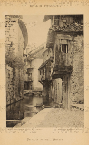 Un coin du vieil Annecy, View of a street on a water in Annecy, page, photomechanical print, maker: F. Thévoz & Cie., maker: Eugène Demole, c. 1887 - in or before 1892, paper, collotype, photoprint, height 159 mm x width 111 mm, street, canals, waters (in city), A sepia-toned photograph of a narrow canal lined by old stone and wooden buildings, with overhanging upper stories and tiled roofs; a small arched bridge spans the canal in the midground; the water reflects the buildings and a simple wooden platform sits at the near edge; a textured stone wall runs along the left side and a cobbled walkway is visible on the right; balconies and small windows appear on the buildings, and wooden beams and shutters show weathered surfaces; decorative printed headings and captions appear on the page surrounding the photograph.