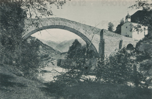 Pont de Neubrücke - Vallée de la Viège, View of the bridge of Neubrück, page, photomechanical print, maker: Frédéric Thévoz, maker: F. Thévoz & Cie., c. 1888 - in or before 1893, paper, collotype, photoprint, height 118 mm x width 174 mm, mountains, bridge, Switzerland, A landscape photograph showing a stone arch bridge spanning a river with vegetation in the foreground and on the riverbanks, leafy branches framing the top of the scene, a steep tree-covered slope to the left, and a white building with a small tower situated near the bridge on the right side, with distant mountain peaks visible through the arch.