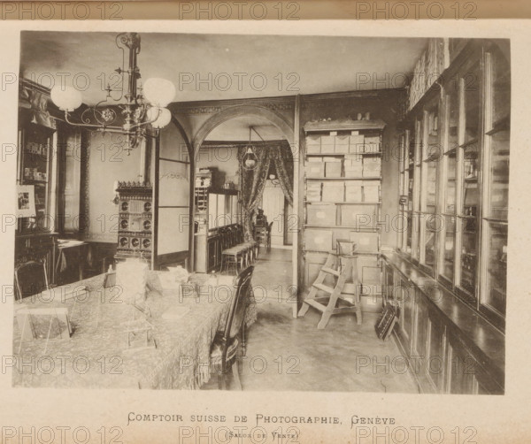 Comptoir suisse de photographie - Salon de Yente, Interior of a Swiss photography shop, page, photomechanical print, maker: F. Thévoz & Cie., c. 1888 - in or before 1893, paper, collotype, photoprint, height 114 mm x width 151 mm, Print on second and third page after page 131., lighting, lamps, edifice, eg: public building, department store - AA - civic architecture: inside, A sepia-toned interior scene shows a room with a long table covered by a patterned cloth and fringed edge, chairs pushed around it, and small display stands and objects arranged on the table. A large ornate chandelier with globe lamps hangs from the ceiling near a decorative stove with ornate panels. An arched doorway divides the space, revealing a curtained area with a hanging lamp and a row of stacked chairs and a small table or desk beyond. To the right, tall shelving holds numerous stacked boxes and a wooden step ladder leaning against them, and a wall of glass-fronted cabinets or windows runs along the far rig...