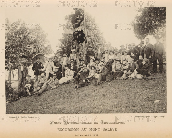 Union Internationale de Photographie - Excursion au Mont Salève, Group portrait of members of the Union Internationale de Photographie, page, photomechanical print, maker: Jules Royer, maker: E. Batault, 1893-08-20, paper, collotype, photoprint, height 105 mm x width 160 mm, anonymous historical persons depicted in a group, in a group-portrait, Salève, Mont, A sepia-toned photograph shows a large group of men and women gathered outdoors on a grassy slope in front of a large tree. The people are arranged sitting, reclining, and standing, many wearing period clothing including suits, hats, and long dresses; several women wear wide-brimmed hats and some men wear bowler or brimmed hats. A few people hold umbrellas, canes, or walking sticks, and one person appears to be seated on a bench at the right edge. Trees and foliage form the background, and the ground in the foreground is grassy with scattered leaves. The image has a wide white margin around it.