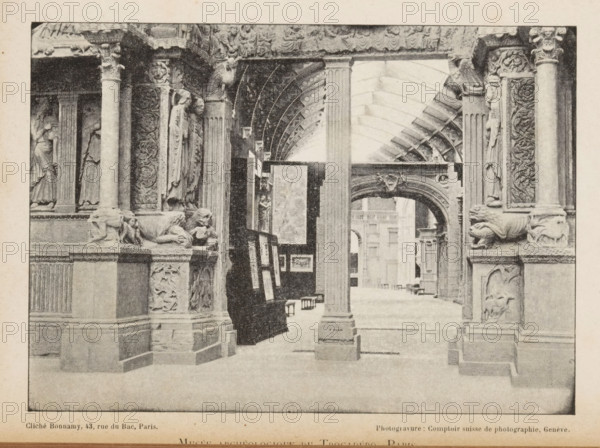 Musée archeologique du trocadéro, Paris, View of an entrance to a room of the Musée archeologique du trocadéro, page, photomechanical print, workshop stamp: 'No 7887, post, tenebras, spero, lucem', cliché maker: Bonnamy, maker: Comptoir Suisse de Photography, c. 1889 - in or before 1894, Comptoir Suisse de Photographie, paper, autotype, height 123 mm x width 173 mm, column, pillar, architecture, exhibition, art, A sepia-toned indoor scene shows a vaulted gallery with ornate stone columns and carved architectural details. Sculpted figures are set into niches and decorative relief panels run vertically along the columns. Stylized animal sculptures rest on ledges near the column bases. An archway with carved ornamentation frames a view into a distant interior space with additional columns and architectural elements. Framed artworks are displayed on easels and leaning against a dark partition beneath the vaulted ceiling. The floor appears smooth and open beneath the sequence of arches and ...