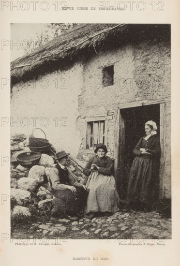Two unknown women and an unknown man in front of a building, Causette du soir, page, photomechanical print, maker: M. Bovagne, maker: Jules Royer, c. 1890 - in or before 1895, paper, collotype, photoprint, height 163 mm x width 122 mm, adult woman, adult man, open door, A black and white photograph shows part of a rustic building with a thatched roof and a textured wall, a small window with wooden framing, and a doorway with a wooden lintel. In front of the building are three people: a seated man wearing a hat and vest, a seated older woman in a dress and apron holding her hands together, and a standing woman in a long dress and headscarf leaning in the doorway with her arms crossed. To the left of the seated man there are woven baskets placed on a pile of stones and some scattered rocks and cobbles on the ground in front of the group.
