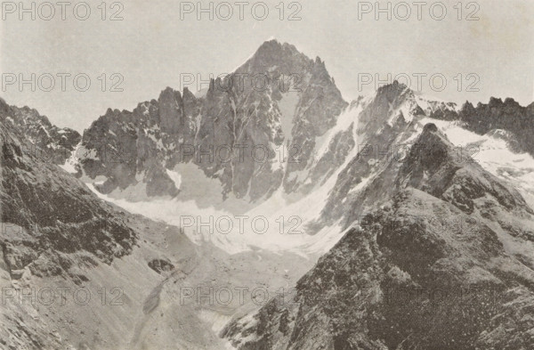 View of the snowy peaks of mountains in Ecrins, Col et dome de neiges des Ecrins, page, photomechanical print, inscription: '(Extrait de l'Echo des Alpes, Geneve.', photographer: ME Courvoisier, c. 1890 - in or before 1895, ME Courvoisier, photographer, paper, autotypie, height 109 mm x width 168 mm, snow, mountains, Écrins, Les, A wide grayscale photograph showing a rugged mountain range with steep jagged peaks and snow-filled gullies, with a prominent central peak rising above surrounding ridges. In the foreground there are rocky slopes and a ridge descending toward a valley floor that appears to be partially covered in snow or ice. The sky above the mountains is clear and light in tone. The image has a textured, grainy appearance.