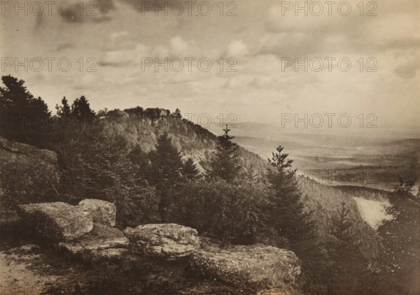 View of the landscape of Odiliënberg, La montagne de Sainte-Odile, page, photomechanical print, maker: Victor Franck, maker: Jules Royer, c. 1890 - in or before 1895, paper, collotype, photogravure, height 121 mm x width 171 mm, trees, mountains, Vosges, Mont Sainte-Odile, A sepia-toned landscape photograph showing a rocky foreground with large flat stones, a densely wooded hillside with coniferous and broadleaf trees rising toward a distant ridge where a cluster of buildings sits, a lone tree silhouette near the ridge edge, and a wide sky filled with soft, layered clouds above a faint, hazy valley stretching into the distance.