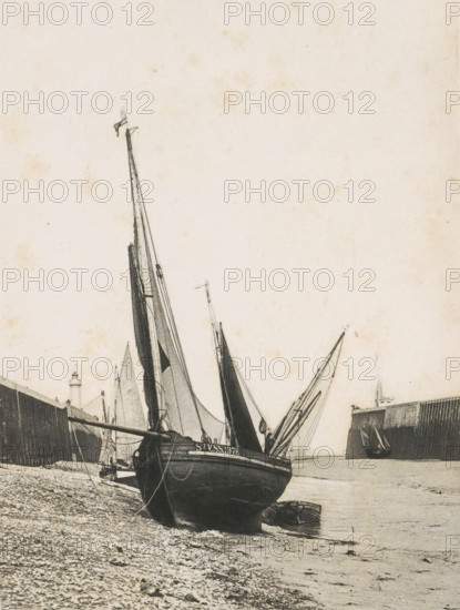 Marée Basse, View of a sailboat stranded at low tide, page, photomechanical print, photographer: Charrel, cliché maker: Dujardin, printer (person): Eudes et Chassepot, c. 1890 - in or before 1895, printer (person), paper, photoaquatint, height 168 mm x width 123 mm, sailing-ship, sailing-boat, ebb, low tide, A monochrome scene showing a large sailboat resting on a pebbled shore with its hull facing the viewer and tall masts rising upward, rigging and sails furled against the spars, a smaller boat pulled up beside it, a long wooden breakwater or pier extending from the left into the water, a lighthouse structure visible near the pier, calm water along the shoreline, another vessel partly visible near the end of a second pier on the right, and a wide expanse of pale sky above.