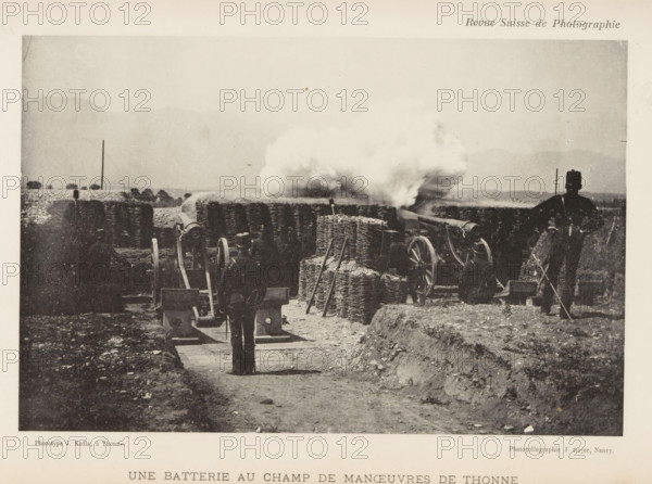 Une Batterie au Champ de Maneuvers de Thonne, Two gunners at Thonne, France, page, photomechanical print, maker: J. Kolla, maker: Jules Royer, c. 1891 - in or before 1896, paper, collotype, photoprint, height 113 mm x width 173 mm, firearms: cannon, Thonne, A grainy black and white scene showing a dirt path flanked by low earthworks and scattered rocks leading toward a line of artillery pieces behind woven wicker or gabion barriers, with plumes of smoke billowing from one of the guns; several uniformed figures stand or kneel near the guns, one figure facing the camera on the path and another standing to the right with a long object in hand; wooden wheels and gun carriages are visible among stacked wicker bundles and upright poles, and distant low hills or mountains form a pale backdrop under a wide sky.
